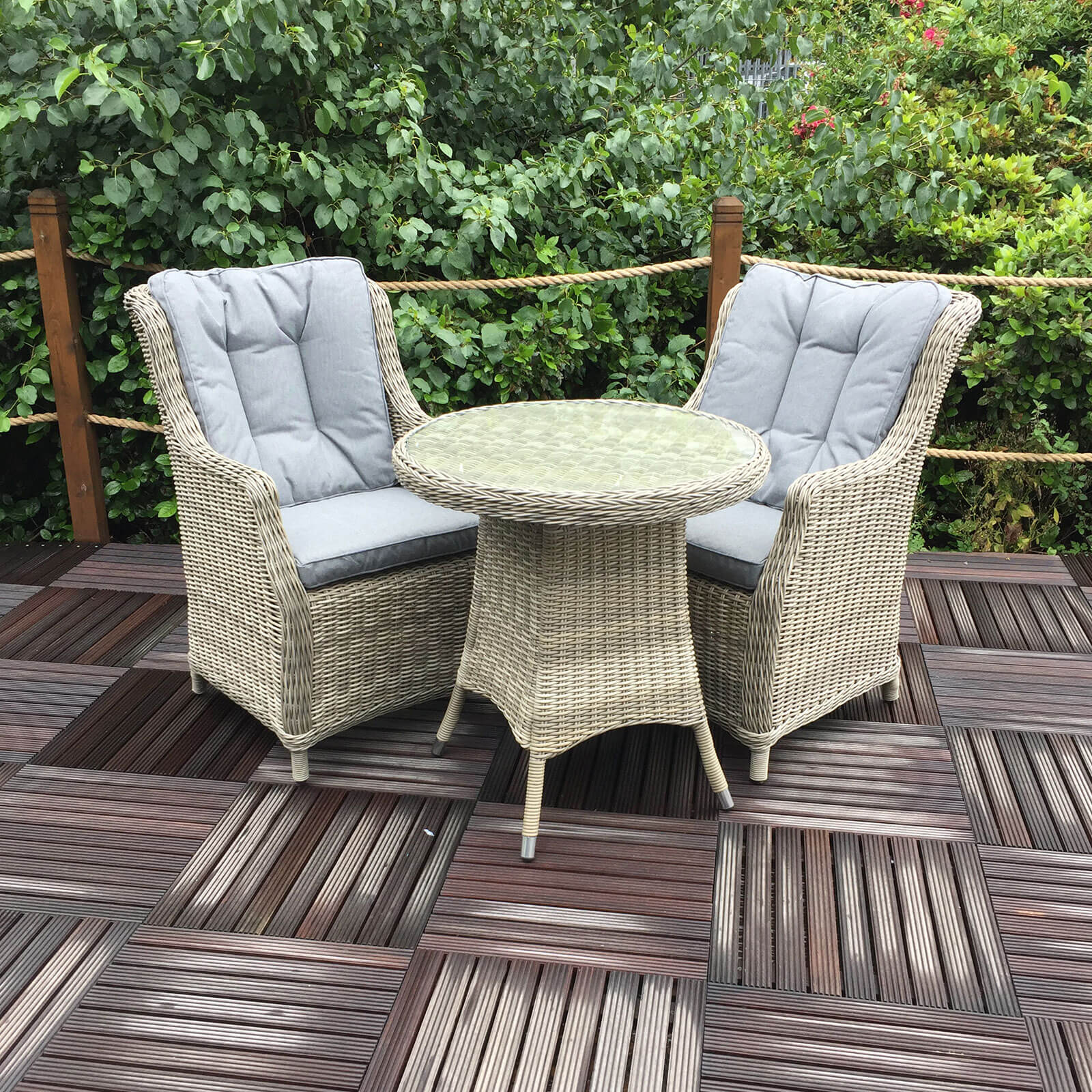 Ballintoy 2 seat round bistro set for the garden or patio area with 2 highback armchairs and round bistro table in beige rattan and grey cushions in a garden setting
